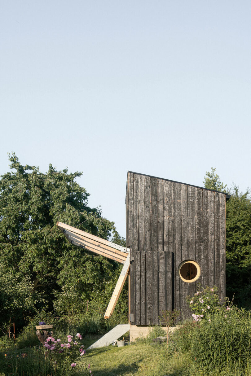 A small, modern wooden cabin with a circular window, situated in a garden with greenery and flowers, and featuring a unique fold-up wall.