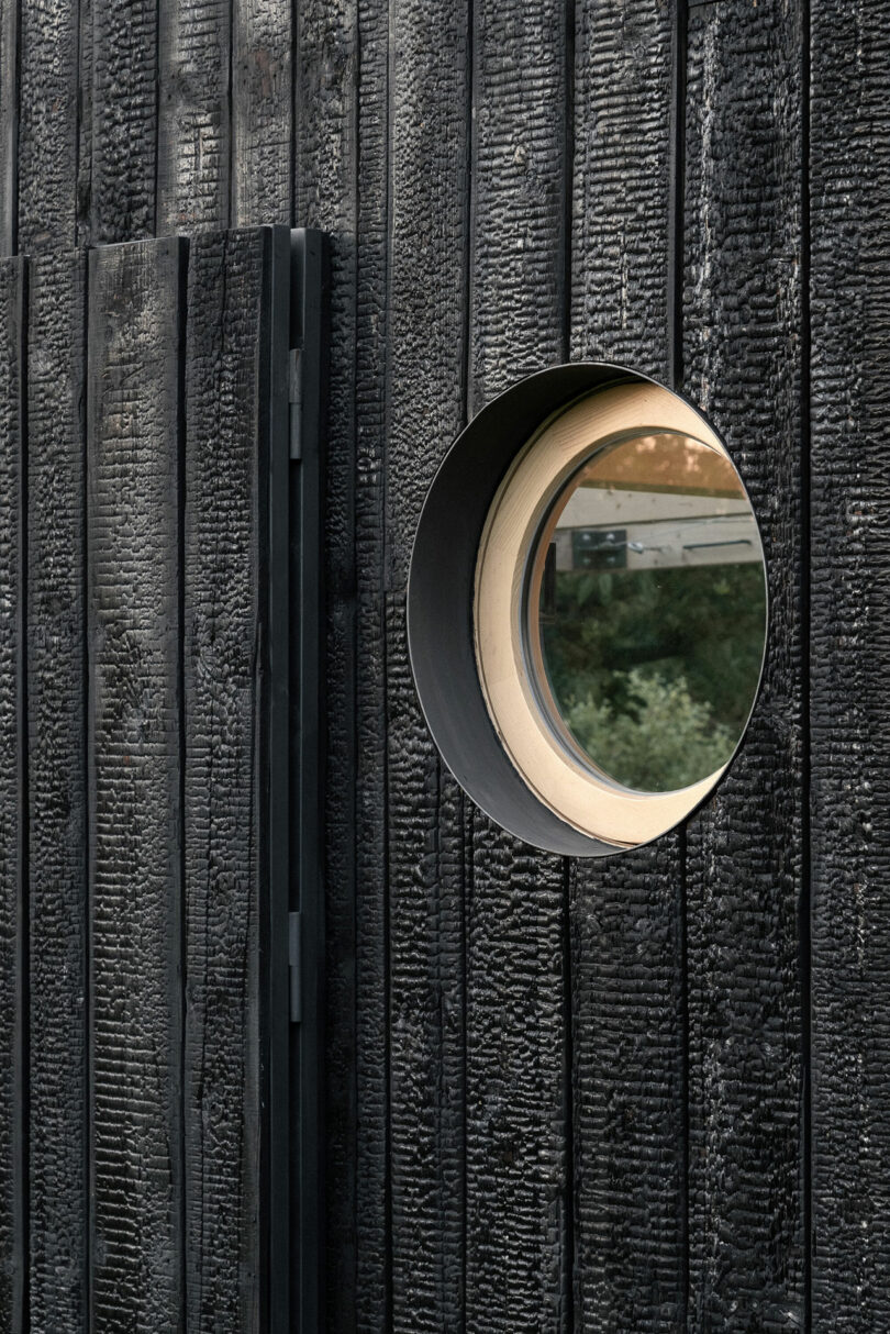 A round window set in a wall of charred black wooden planks with a partial reflection of greenery visible in the glass.