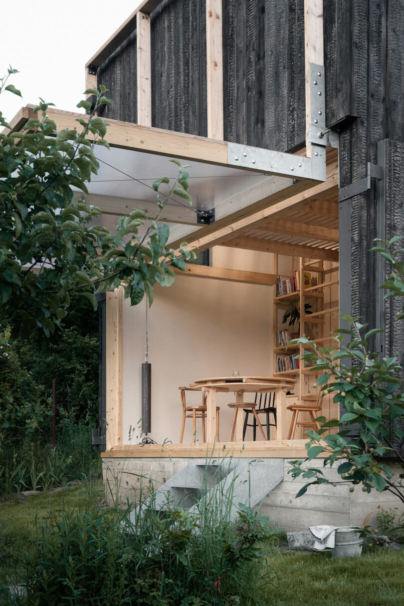 Outdoor view of a modern wooden building with an open door leading to a cozy interior with a dining table and chairs, surrounded by lush greenery and garden plants.