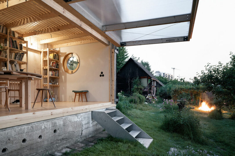 A modern wooden house with an open front showcasing its interior, including shelves and furniture. Steps lead to a green yard with a garden, trees, and a small fire pit in the background.