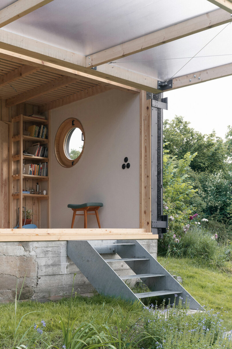 A small wooden structure with an open side revealing a cozy interior. There is a circular window, a bookshelf, a stool, and a metal staircase leading to the grassy outdoors.