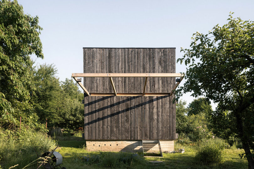 A small, dark wooden cabin with a modern design stands in a grassy, forested area, featuring a simple rectangular shape and exposed beams.