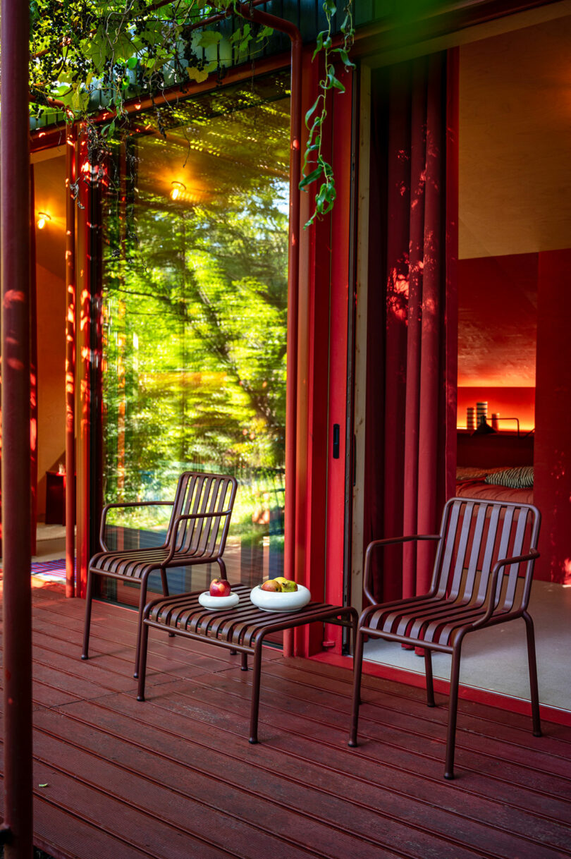 A small patio with two metal chairs and a table on a red wooden deck outside a glass door, partially covered by greenery.