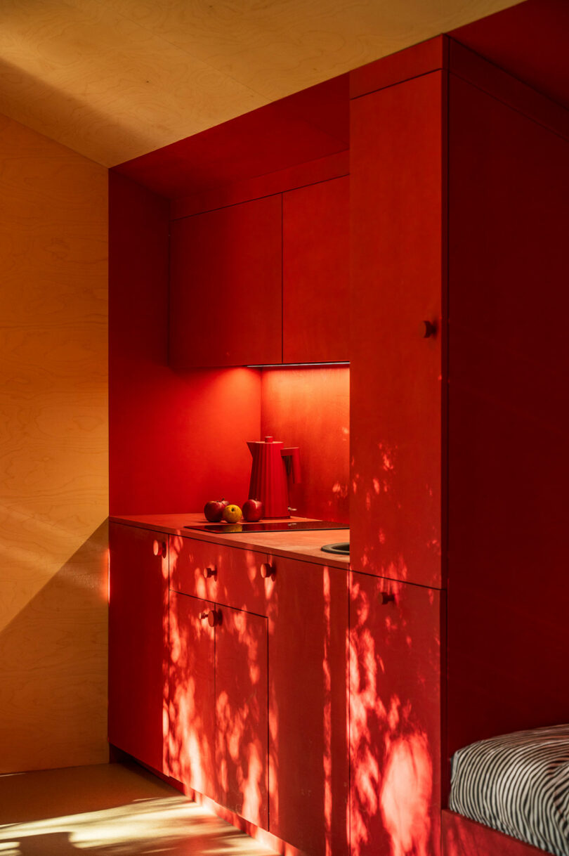 A small kitchenette featuring vibrant red cabinets, a matching red electric kettle, and a few pieces of fruit on the counter. Sunlight casts tree shadows on the cabinets, adding texture to the red surfaces.