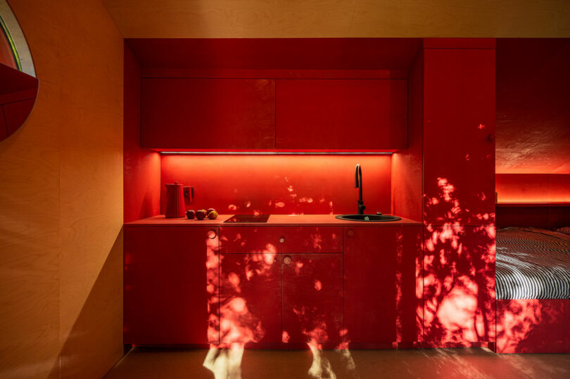 A minimalist red kitchen with a round sink, faucet, stovetop, and kettle. Sunlight filters through trees, casting dappled shadows on the cabinets and counter.