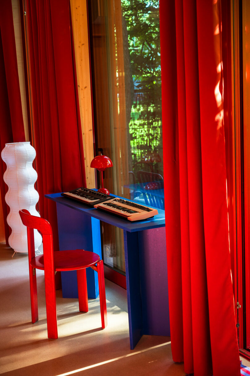 A room with red curtains features a red chair, a blue desk with a keyboard, and a red lamp. A white, wavy floor lamp stands nearby. Natural light streams in from the windows.