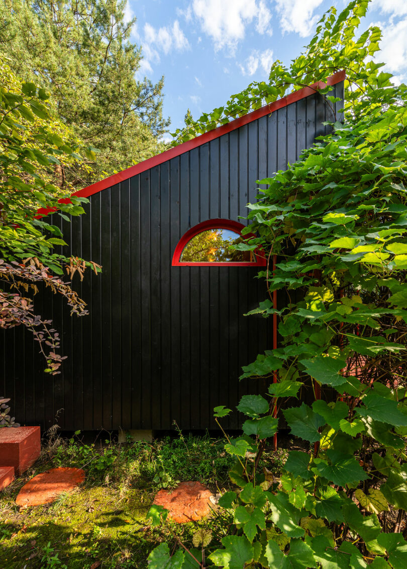 A small black wooden building with a slanted roof and red accents is surrounded by lush greenery, including trees and plants.