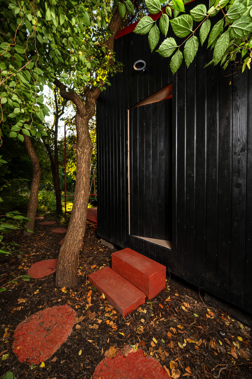 A small black wooden building with a red door and steps is surrounded by lush green trees and shrubbery. Red stepping stones lead to the entrance.