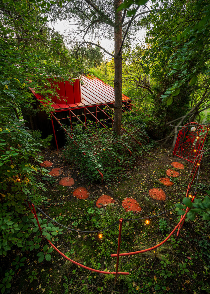 A small garden shed with a red roof is surrounded by lush greenery. It features a red metal gate and stepping stones leading up to it through the overgrown garden.