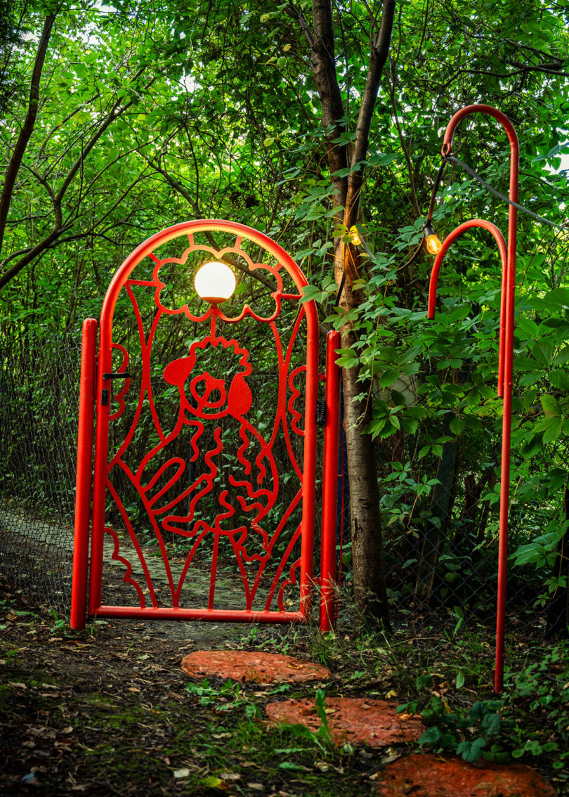 A red metal gate with an ornate design stands beside a curved red lamp post in a lush, wooded area.