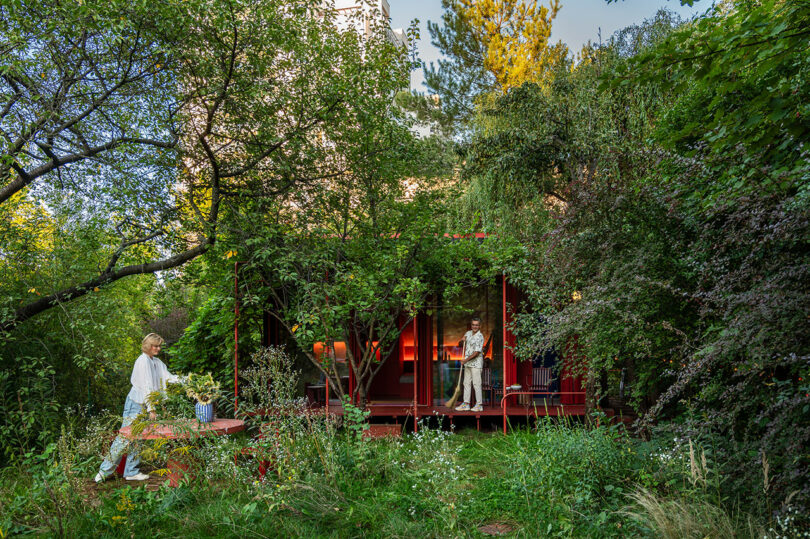 A person arranges flowers on a table while another person works on a laptop on a porch surrounded by lush green trees and plants.