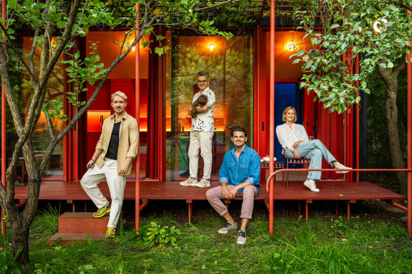 Four people are posing on the porch of a red house surrounded by greenery; one person is holding a small dog.