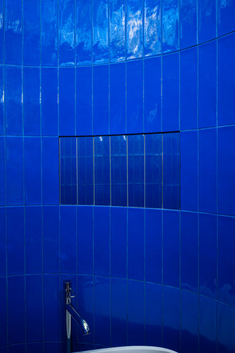 A wall covered in shiny blue tiles features a rectangular niche with smaller, darker blue tiles. A modern faucet is partially visible at the bottom of the image.