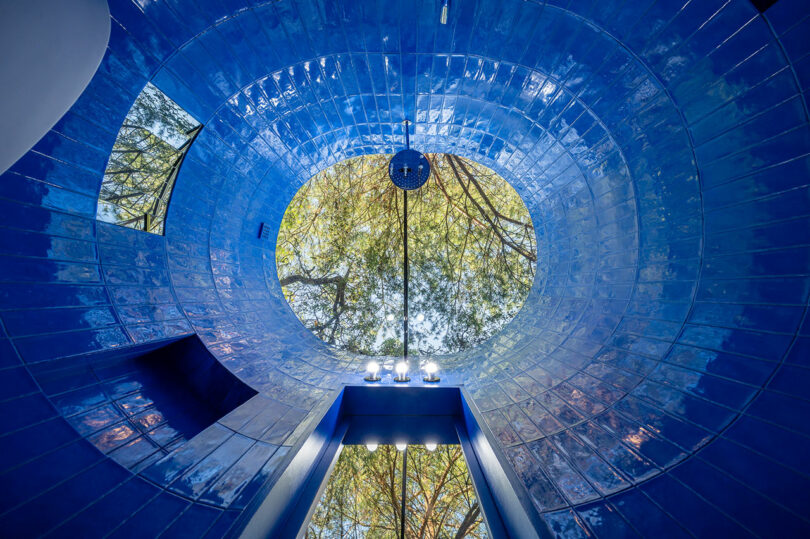 Blue-tiled room with a circular skylight opening to a view of trees. The skylight frames branches and leaves, creating a natural contrast with the vivid blue tiles.