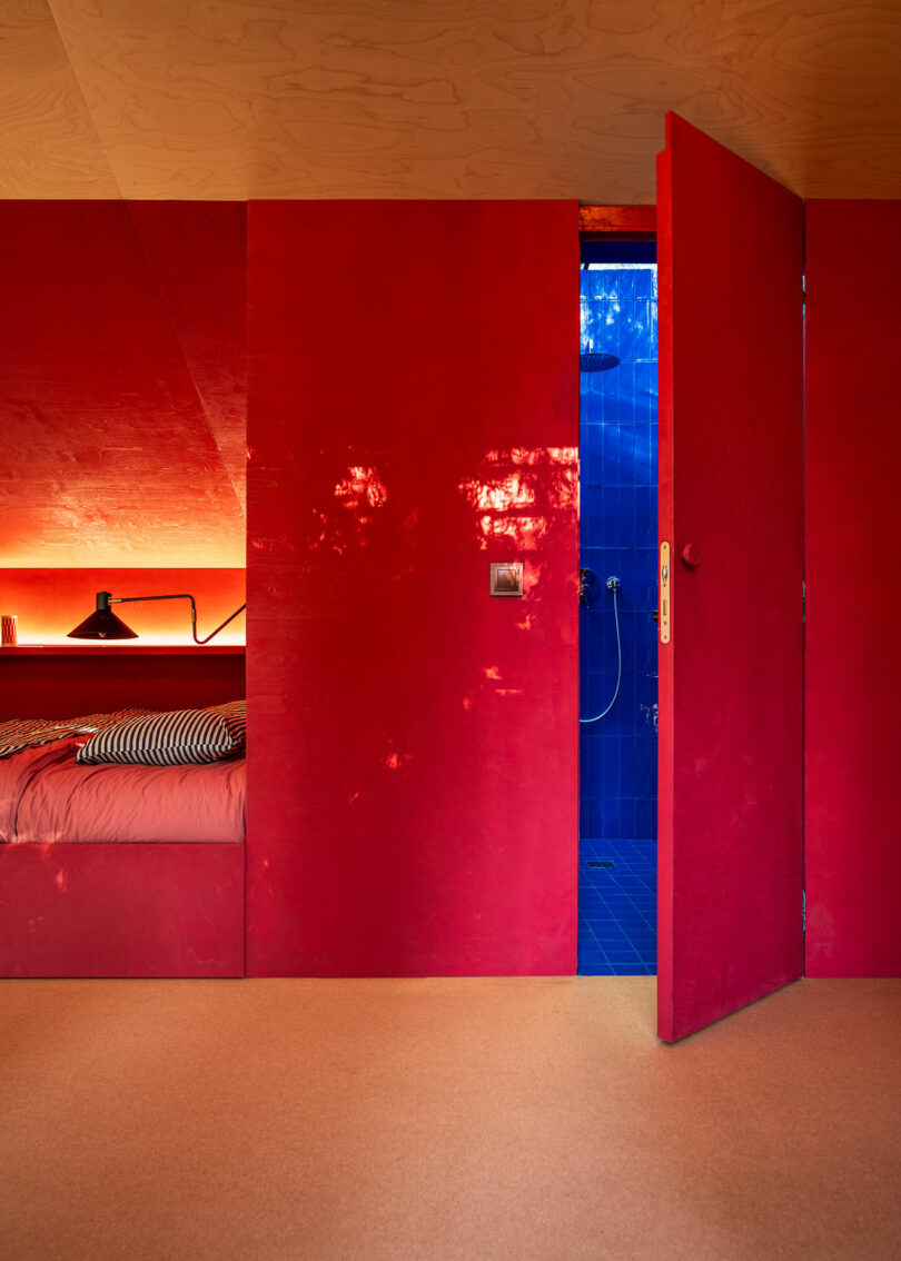 A room with red walls and a partially open door revealing a blue-tiled bathroom. There are shelves with lights and a bed on the left side, and a black lamp on the shelf.