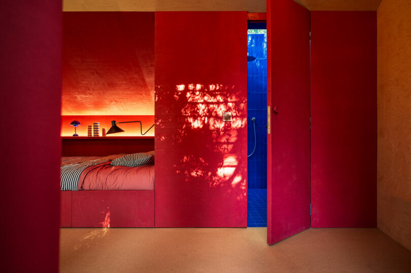 A bedroom with vibrant red walls and a partially open door revealing a blue-tile room. The bed is made with striped linens, and a desk lamp and small items are on the headboard shelf.