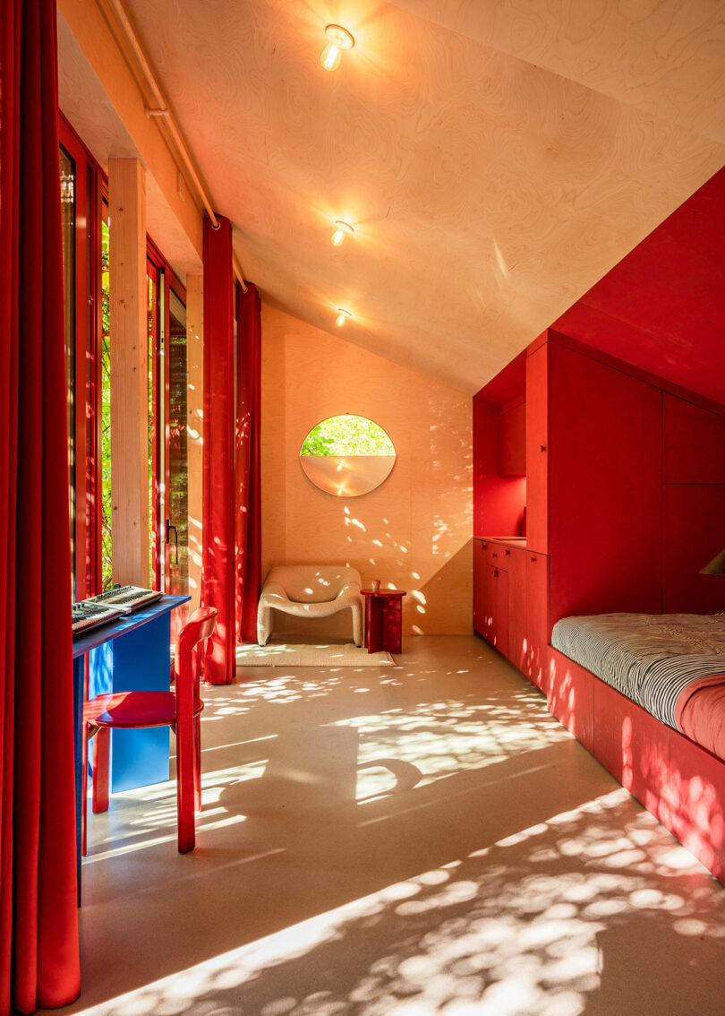 A colorful room with vibrant red walls, a small desk and chair, a window with draped red curtains, and a bed. Sunlight filters through the windows, casting dappled shadows on the floor and ceiling.