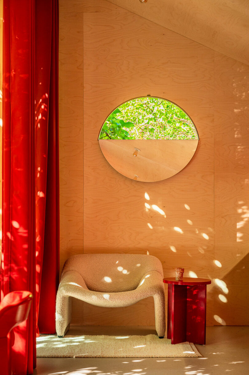 Room with wooden walls, a round window with greenery outside, a white armchair, red curtains, and a red side table with a glass on it.