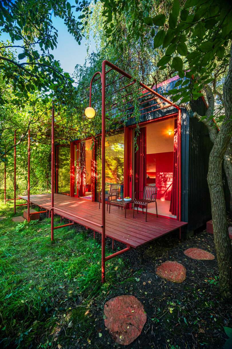 A red wooden cabin with a glass door, a patio with two chairs and a table, surrounded by lush greenery and trees, illuminated by a warm outdoor light.