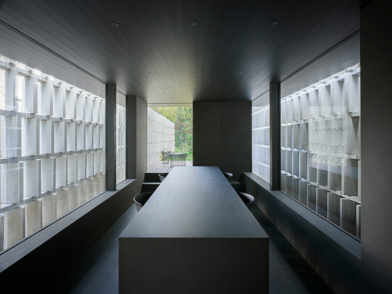 A modern, minimalist dining room with a long rectangular table, surrounded by gray walls and large windows with geometric patterns, allowing natural light to filter through.