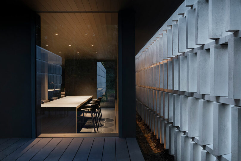 A modern dining room with large glass windows, a long table, and several black chairs is adjacent to an exterior wall with a geometric grid design.