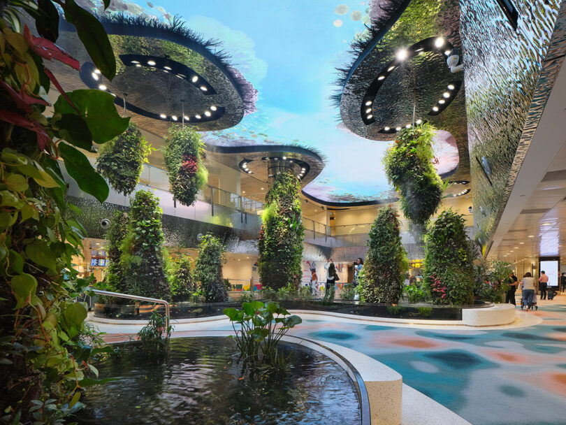 A modern indoor space with abundant plant life, water features, and a unique ceiling design resembling an underwater scene. People are walking and interacting in the area.