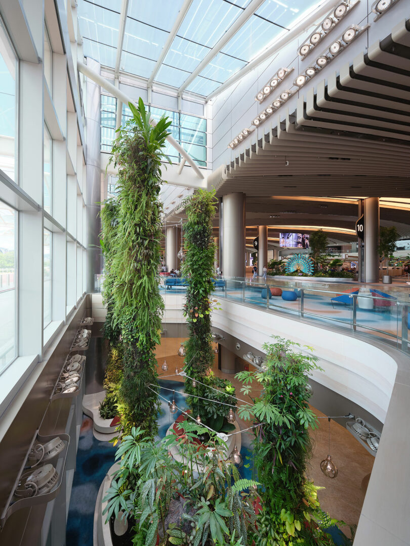 Indoor garden with tall, vertical plant installations and various tropical plants under a glass ceiling, located in a modern, multi-level atrium. A glass railing separates the garden from the walkway.