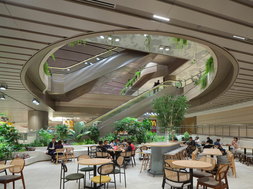 A modern indoor space with a large circular opening in the ceiling, an escalator, and several people sitting at tables surrounded by greenery and plants.