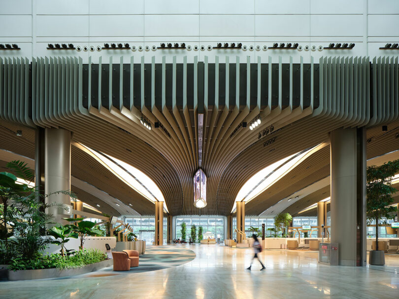 Modern architectural interior with wooden ceiling slats, wide open space, indoor plants, seating areas, and wide windows. Person walking through the center of the image.