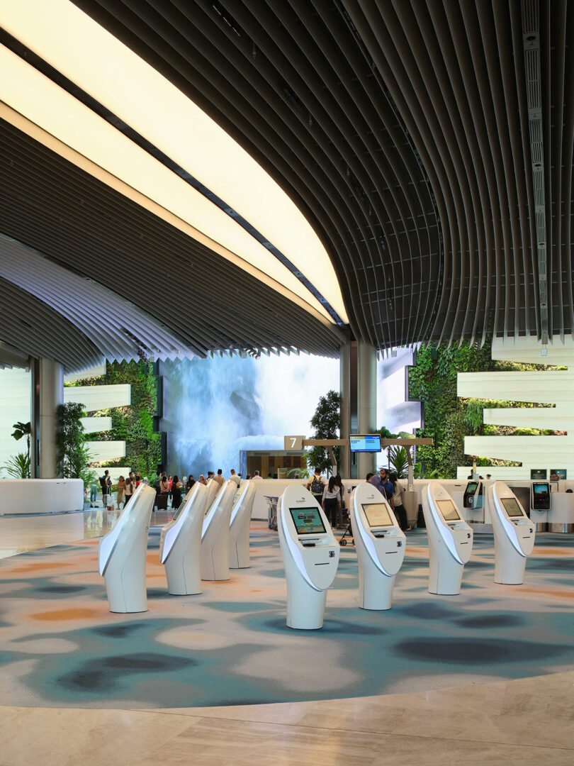 A modern airport terminal with a high ceiling and curved designs, featuring self-service kiosks in the foreground and large digital screens displaying flight information in the background.