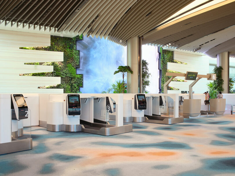 A modern airport check-in area with self-service kiosks, numbered 1 through 6, featuring white counters and a backdrop of a wall with plants and an artistic display.