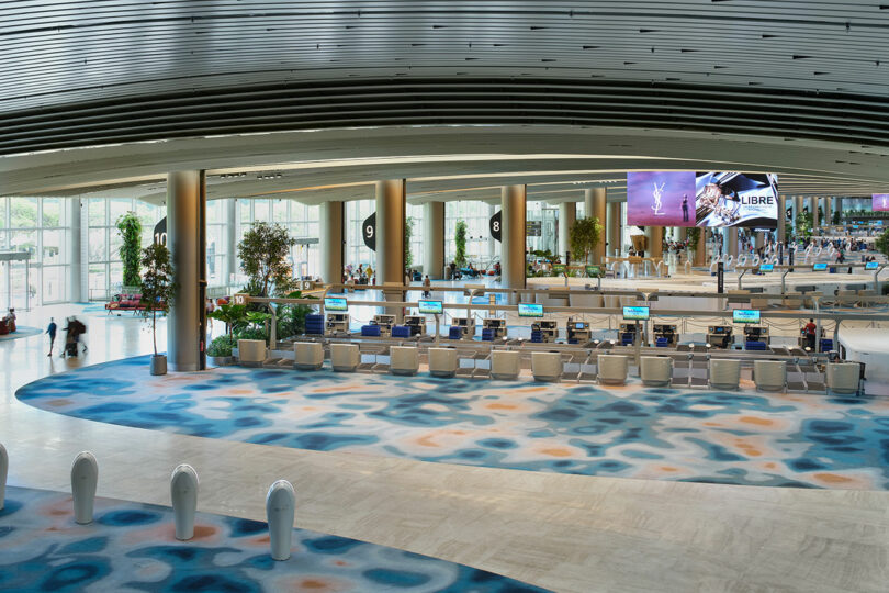 A modern airport terminal with check-in counters, large windows, digital screens, and blue and orange patterned floor. People are visible in the distance.