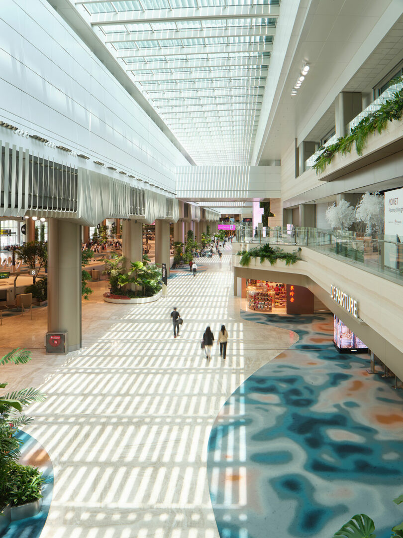 Spacious, modern airport terminal with high glass ceilings, natural light, indoor plants, and a few people walking. The departure area is visible with a kiosk and seating nearby.