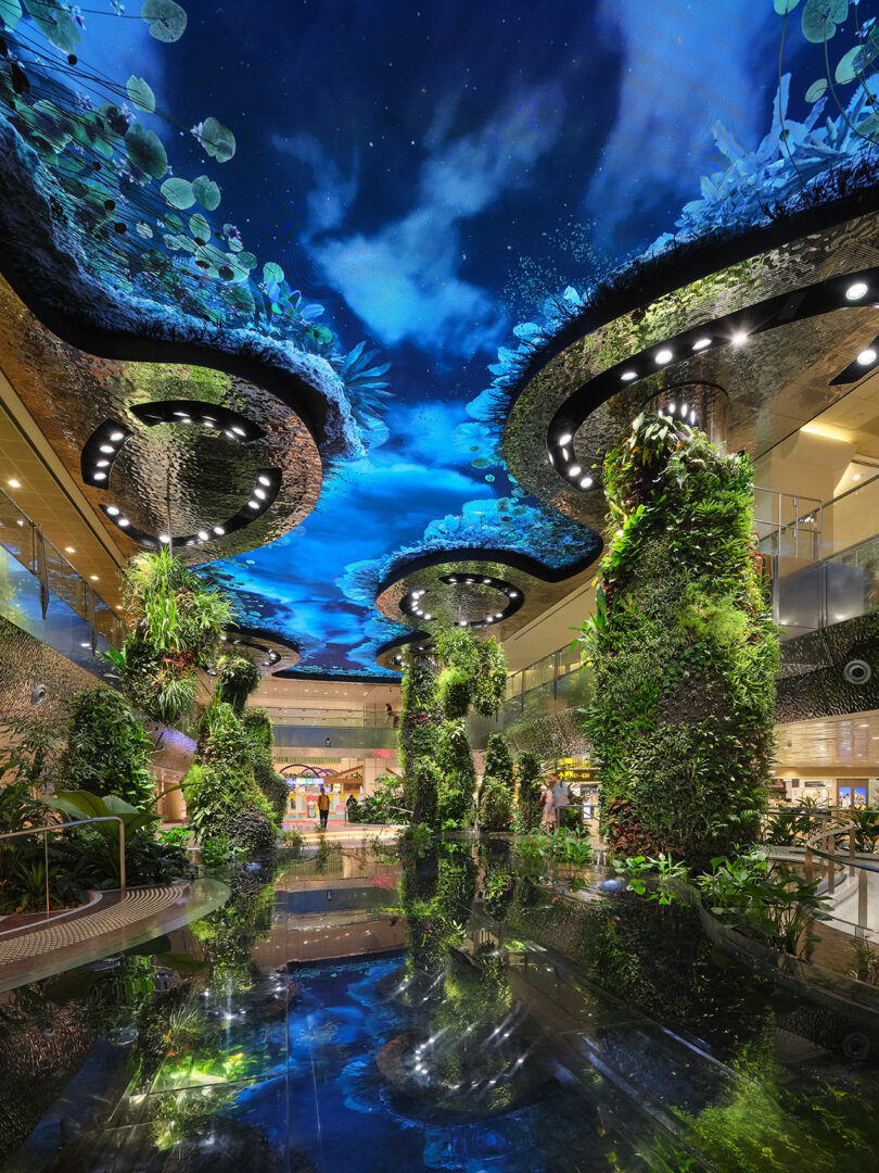 Indoor garden with tall cylindrical plant structures and a ceiling displaying a digital sky and foliage, reflecting off a central water feature.