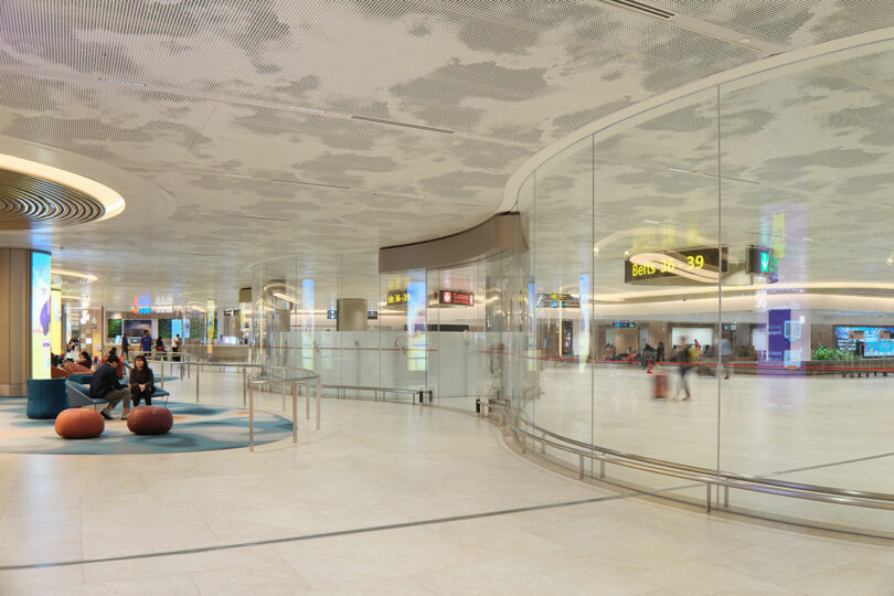 Spacious modern airport terminal with curved glass walls, seating areas, and ceiling lighting. People are seated on cushioned chairs while others walk through the terminal.