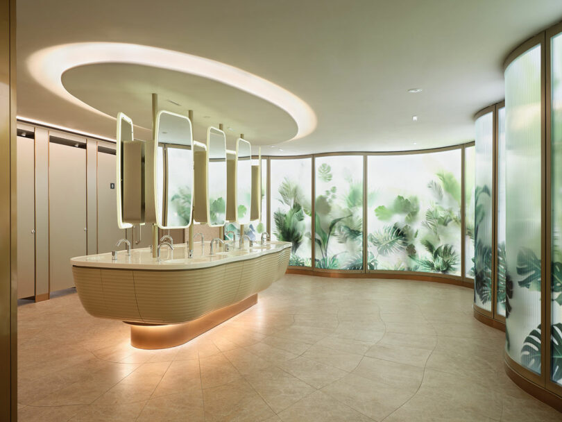 A modern public restroom features a curved vanity with multiple mirrors and sinks, illuminated by ambient lighting. The walls display a serene nature-themed design with frosted glass panels.