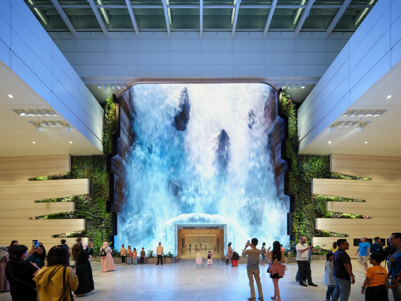 People gather in a spacious, modern lobby featuring a large, illuminated waterfall display. Some are taking photos, while others observe or walk by. The ceiling has a grid-like skylight structure.