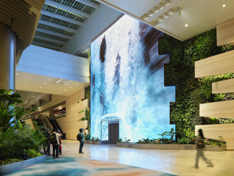 Large indoor waterfall feature with surrounding greenery in a modern, well-lit space. People stand observing the installation. Light-colored walls and plants are part of the decor.