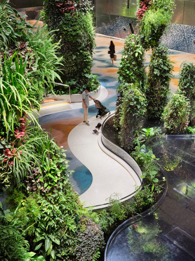 A person stands with a stroller on a winding path surrounded by tall greenery and plants indoors. Another person walks in the background on a colorful floor.