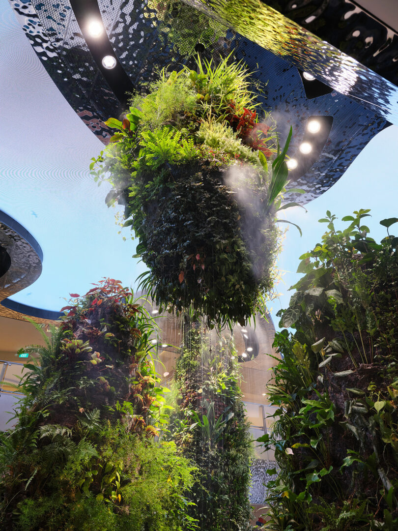 Large cylindrical structures covered in dense green foliage hang from the ceiling, mist being sprayed onto the plants. The background reveals a modern architectural setting with lights and a blue sky.