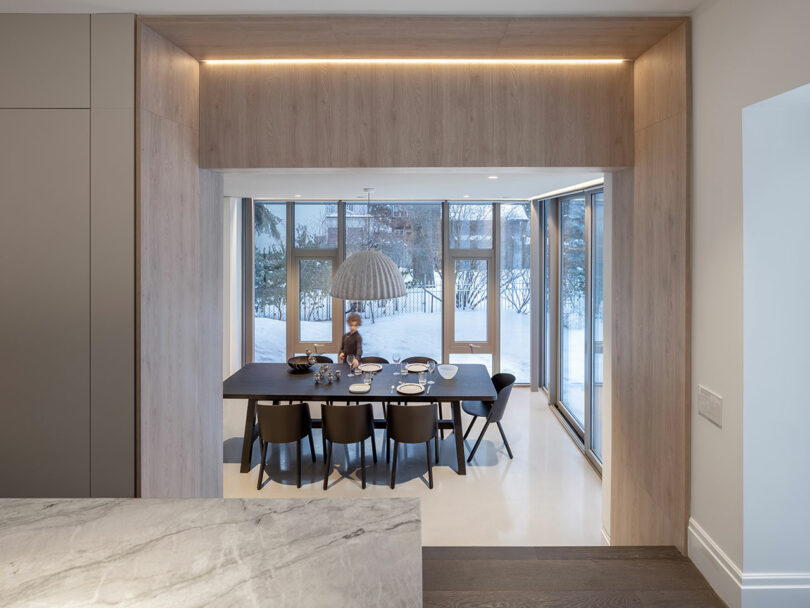 View of a modern dining area with large windows reflecting the architecture, a wooden table set for a meal, and a person sitting at the table. A minimalist light fixture hangs above. Snow is visible outside the windows.