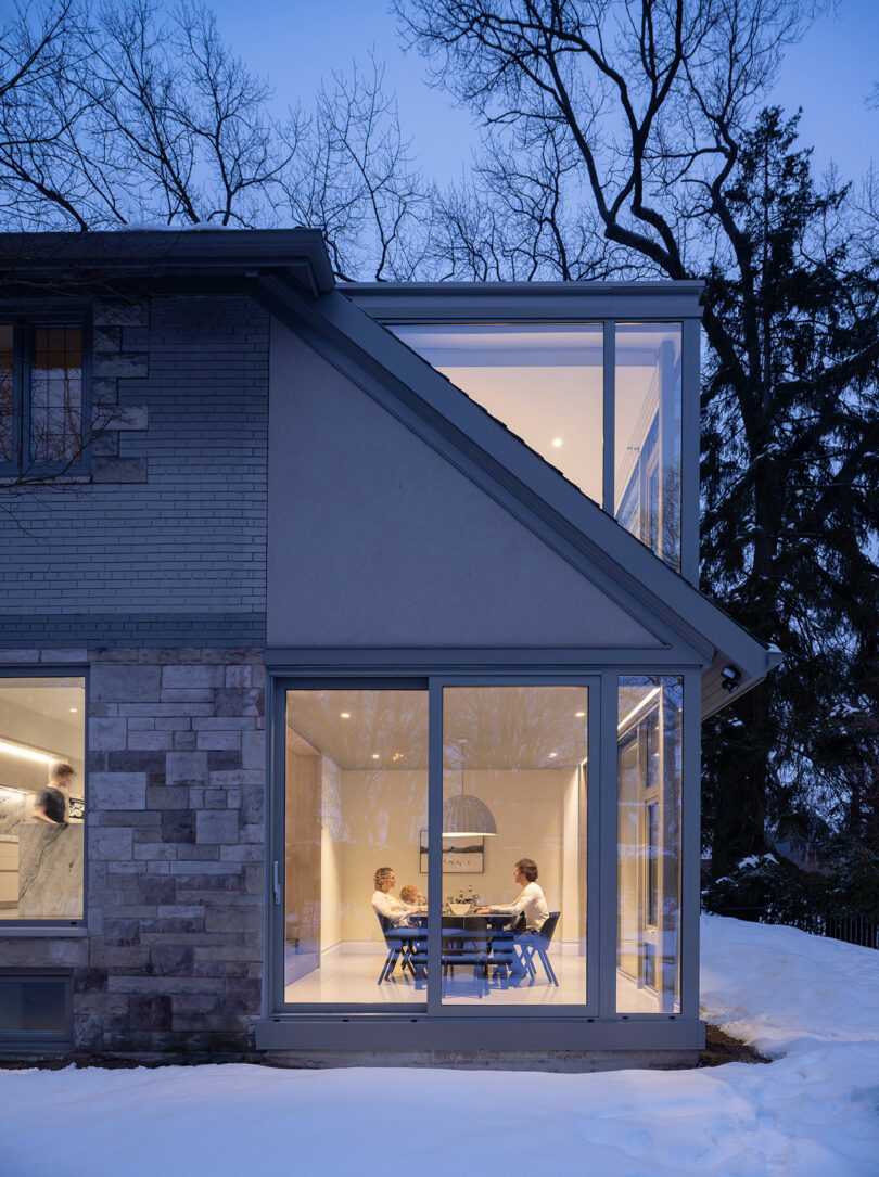 Two people are seated at a dining table inside a modern house with large glass windows, reflecting the sleek architecture. Snow covers the ground outside, and the surrounding trees are bare. The house has brick and glass exterior walls.