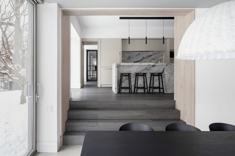 Modern kitchen and dining area reflecting architecture with a marble kitchen island and black bar stools. The space features large windows, dark flooring, and a harmonious mix of light and dark furniture.