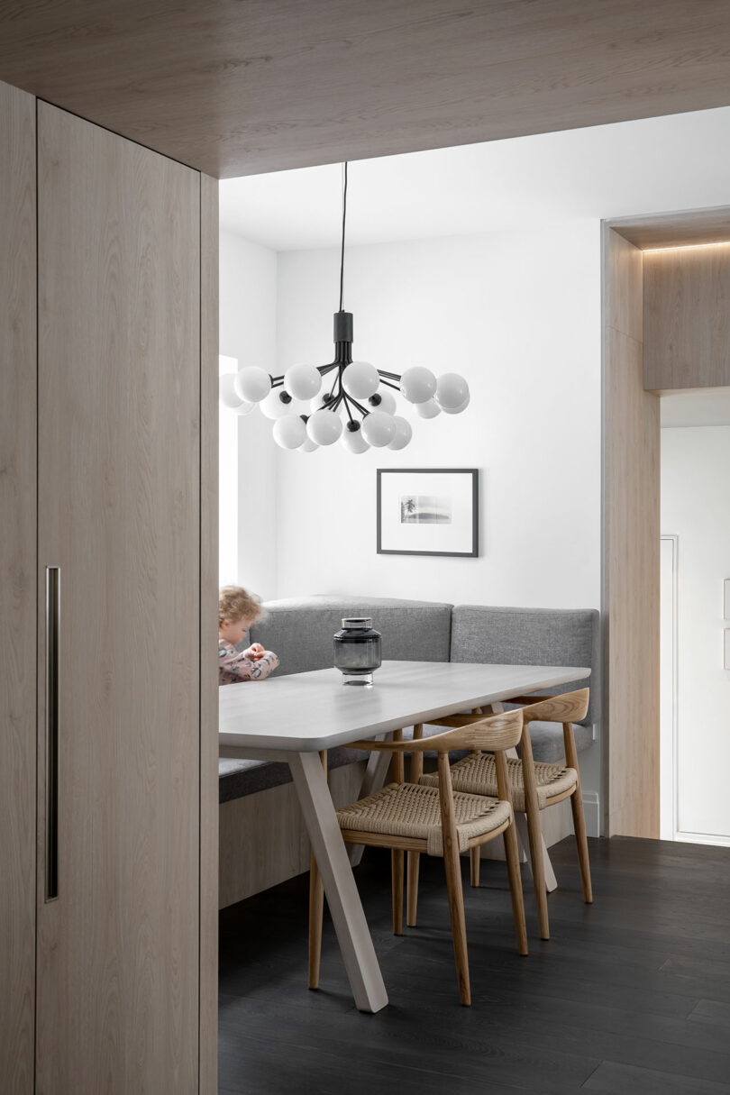 Modern dining nook with a white table, grey cushioned bench, wooden chairs, and a chandelier that reflects architecture. A baby sits on the bench, and a black and white photo decorates the wall above.