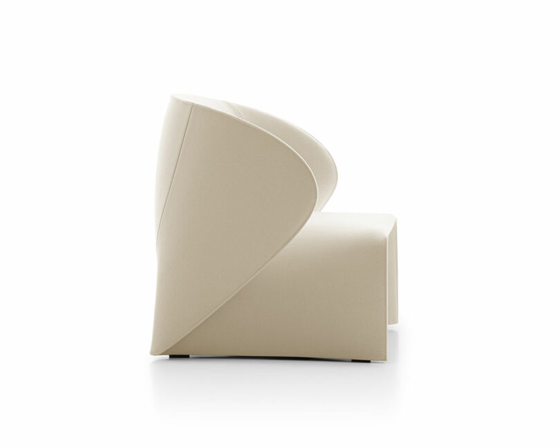 Side view of a modern, beige upholstered chair with a unique, curved backrest design, set against a white background.