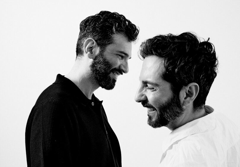 Two men with beards, one in a black shirt and the other in a white shirt, smiling at each other against a plain white background.