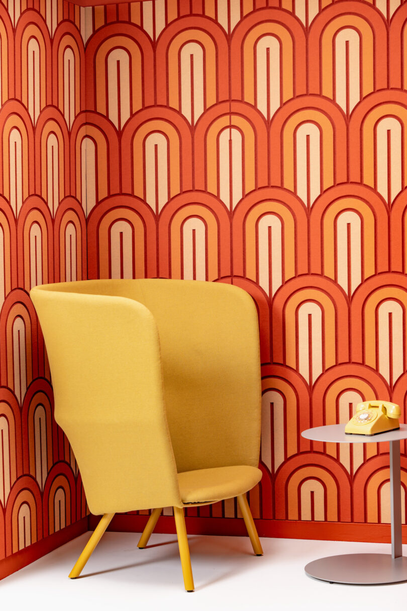 A yellow high-back chair sits in a corner of a room with orange and red patterned wallpaper. A small round table with a yellow rotary phone is positioned next to the chair