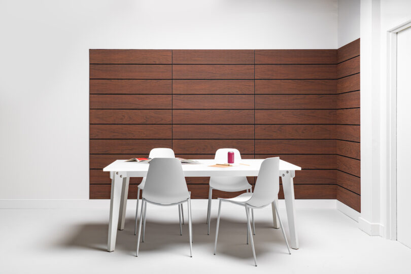 A white table with four white chairs stands in a modern room with wooden paneling on the walls. The table holds a few books and a small decorative object