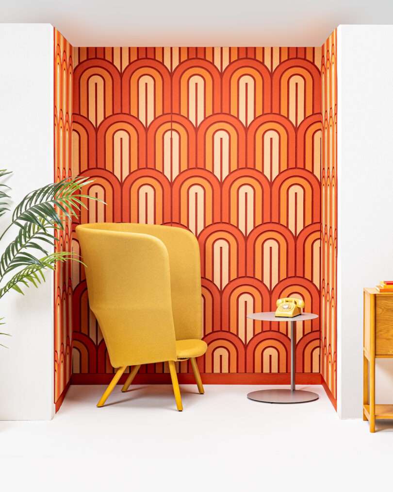 A seating nook with a yellow chair, a small round table, and a vintage yellow rotary phone against a vibrant orange and red retro-patterned wallpaper