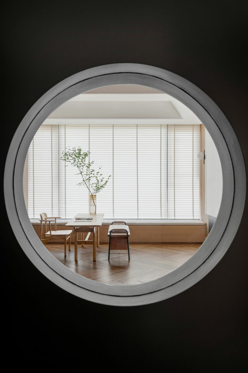 View through a circular opening of a minimalist dining area with a wooden table and chairs, a small tree in a vase, and white blinds covering large windows.